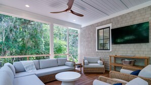 A modern living room with a sectional sofa, a round coffee table, wicker furniture, a flat-screen TV mounted on a brick wall, and large windows overlooking a forested area. - Fabulous 5 BR/5.5 BA Home! New Construction! Pool! Screen Porch! Amenity access included (Seabrook Island)