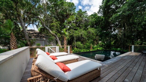 A wooden deck with wicker lounge chairs and white cushions, overlooking a swimming pool surrounded by lush greenery. - Fabulous 5 BR/5.5 BA Home! New Construction! Pool! Screen Porch! Amenity access included (Seabrook Island)