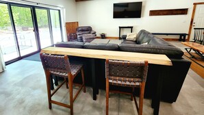A modern living room with a large sofa, two wooden chairs, a wooden table, and a TV mounted on the wall. - Barndominium on H-58 near Pictured Rocks Attractions 
 (Shingleton)