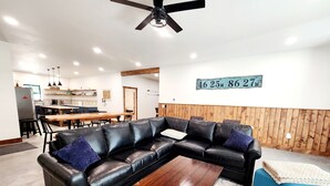 A modern living room with a black leather sectional sofa, a wooden coffee table, and a blue accent chair. A kitchen with a dining area is visible in the background. - Barndominium on H-58 near Pictured Rocks Attractions 
 (Shingleton)