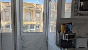 A compact kitchen with a built-in microwave, a coffee machine, and a toaster. A window reveals a balcony with a view of neighboring buildings. - Apartamento Santiago de la Ribera (San Javier)
