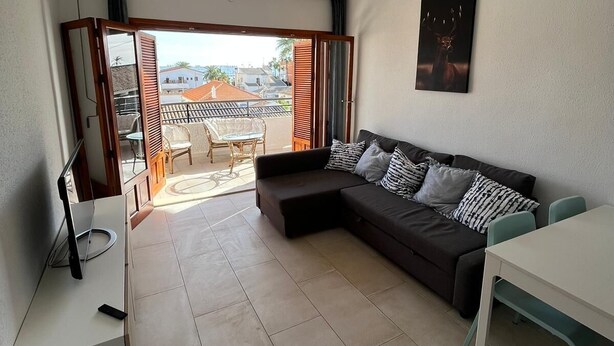 A modern living room with a sectional sofa, a ceiling fan, a dining table, and a view through a glass door to an outdoor area with a patio and chairs. - Apartamento Santiago de la Ribera (San Javier)