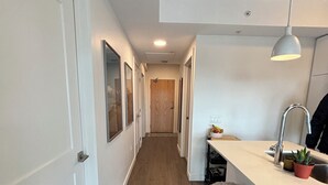 A narrow hallway with wooden flooring, white walls, and a kitchen area with a counter, stools, and a hanging light fixture. - Trendy & Cozy Downtown Halifax Condo (Halifax)