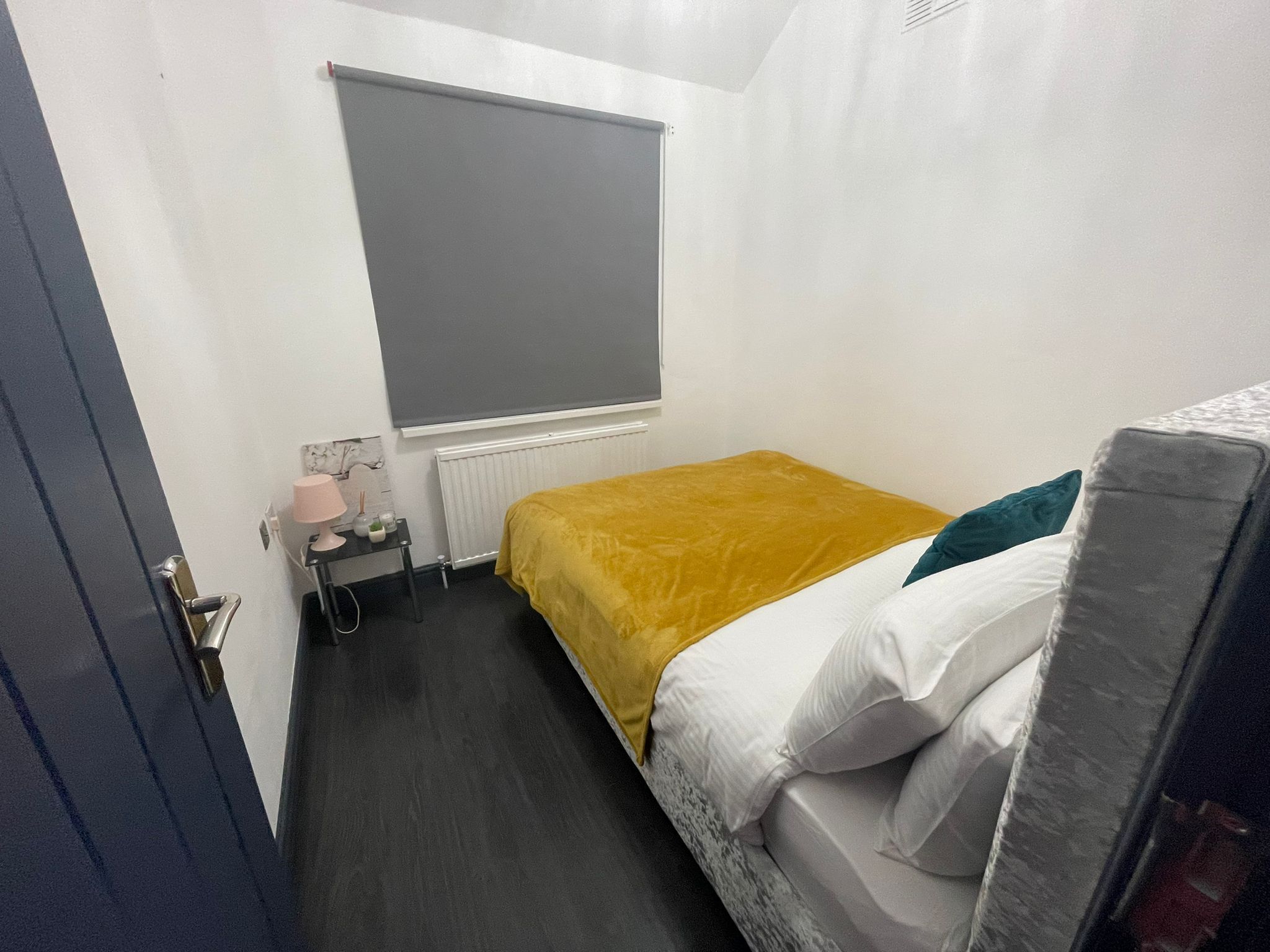 A small, compact room with a bed, a bedside table, a lamp, and a window with a blind.