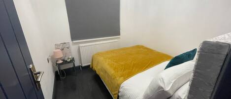 A small, compact room with a bed, a bedside table, a lamp, and a window with a blind.