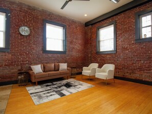 A room with a brick wall, a wooden floor, a brown sofa, two white chairs, and a large window.