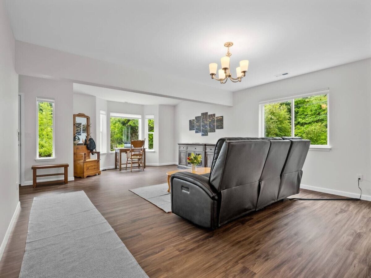 A spacious living room with a dark leather sofa, wooden flooring, and large windows offering a view of greenery.
