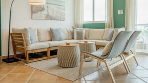 A cozy living room with a sectional sofa, two round side tables, a floor lamp, and a large window with curtains.