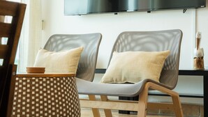 A modern living room with two grey upholstered chairs, a wooden side table, and a checkered ottoman. A flat-screen TV is mounted on the wall above the seating area.