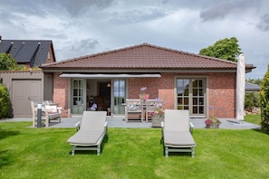 A two-story house with a brick facade, a tiled roof, and a covered patio area with two lounge chairs and a table.
