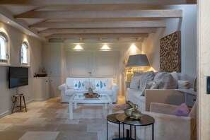 A spacious living room with a white sofa set, a coffee table, and a TV. There is a fireplace with a lamp, and a decorative wall piece.