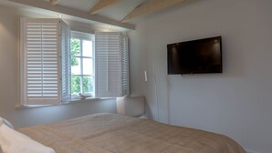 Une chambre à coucher avec un lit, une fenêtre avec des volets, un téléviseur fixé au mur et une petite table sur laquelle est posé un objet décoratif.