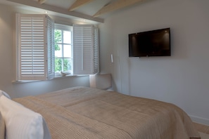 A bedroom with a bed, a window with shutters, a television on the wall, and a small table with a decorative item.