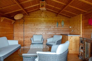 A cozy log cabin interior with a glass-top table surrounded by four wicker chairs, a wooden ceiling, and a rustic wooden cabinet.