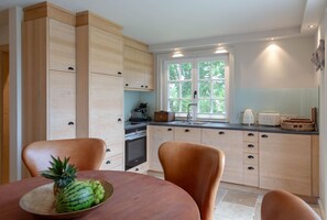 A modern kitchen with wooden cabinets, a dining table with chairs, and a window with a view of trees.
