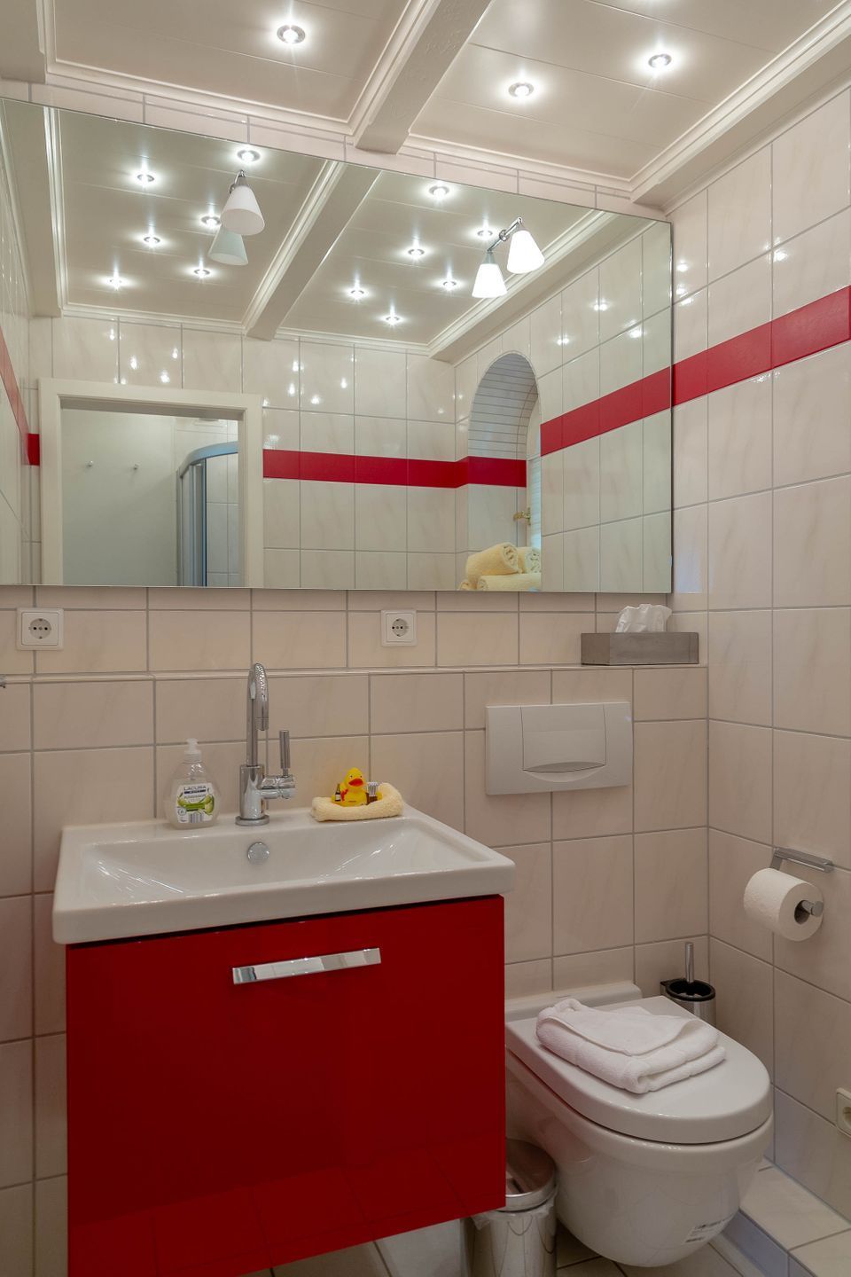 Casa de banho com lavatório branco, armário vermelho e sanita branca. Há um espelho sobre o lavatório e uma faixa vermelha percorre a parede.