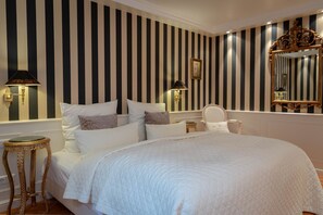 Une chambre d’hôtel avec un grand lit, un papier peint à rayures, deux lampes fixées au mur, un miroir et une petite table.
