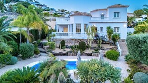 Una villa mediterranea a due piani con piscina, immersa nel verde e palme.