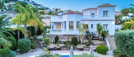 Una villa mediterránea de dos pisos con piscina, rodeada de exuberante vegetación y palmeras.