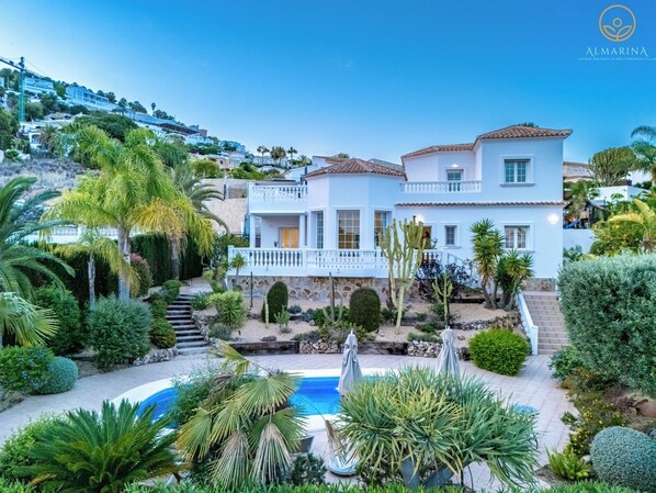 A two-story Mediterranean villa with a pool, surrounded by lush greenery and palm trees.