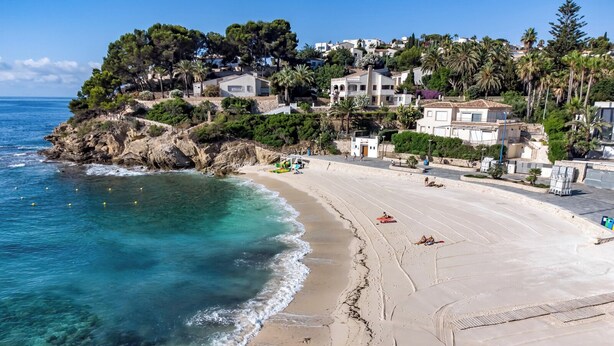 Una spiaggia costiera con acque turchesi trasparenti, una battigia sabbiosa e una collina con edifici residenziali.