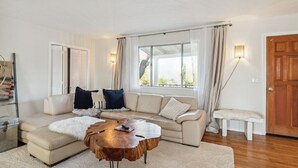 Ein modernes Wohnzimmer mit einem beigefarbenen Ecksofa, einem runden Couchtisch aus Holz und einem großen Fenster mit Vorhängen.