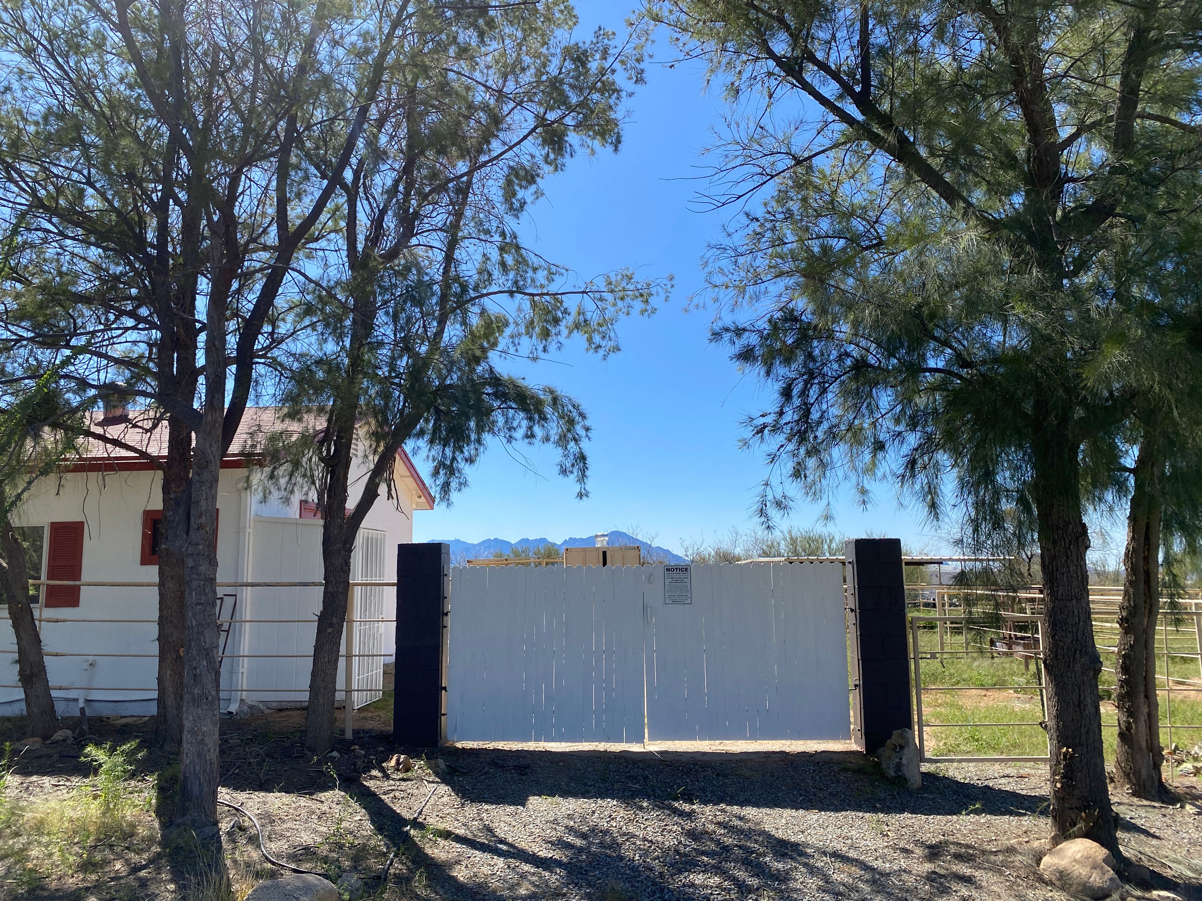 A gated area with a white fence, trees, and a building with red shutters in the background.