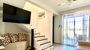 A living room with a sofa, a glass coffee table, a TV mounted on the wall, a staircase, and a sliding glass door leading to an outdoor area.