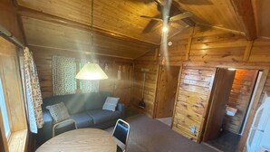 Interior acogedor de una cabaña con techo de madera, ventilador de techo, un sofá, una mesa redonda y una puerta que da a otra habitación.