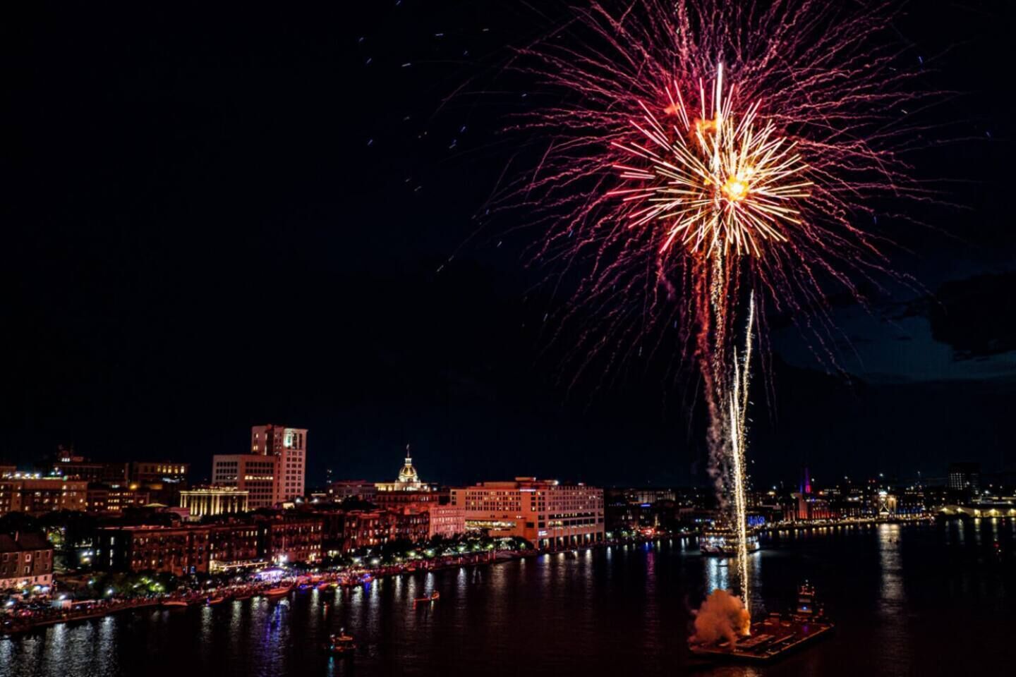 A nighttime cityscape with a prominent fireworks display over a river, buildings along the riverbank, and a boat docked near the waterfront.