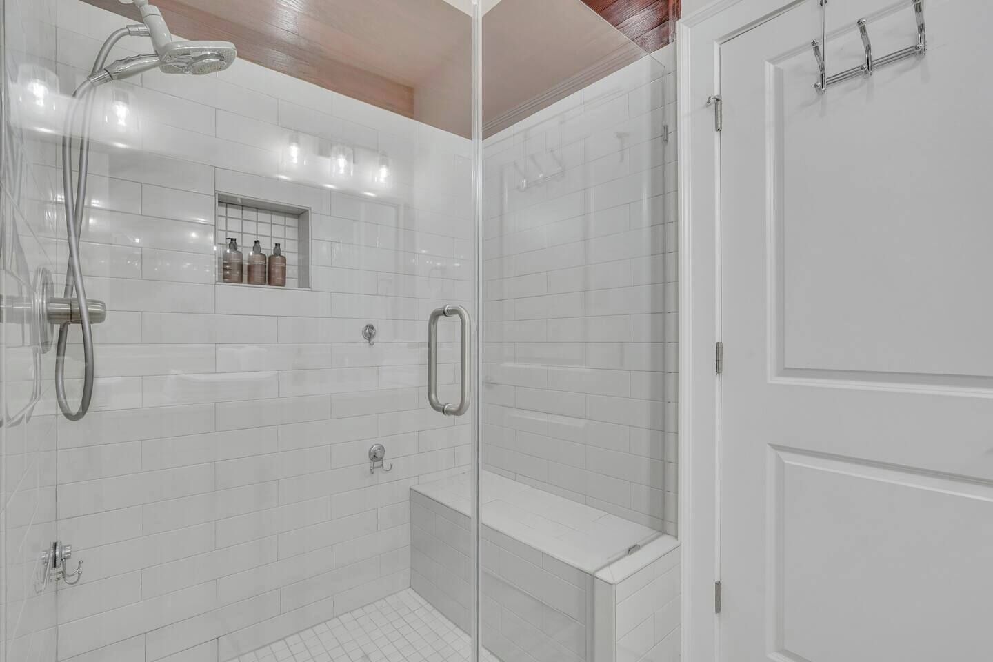 Een moderne badkamer met witte betegelde wanden, een glazen douchecabine, een inbouwkastje en een deur met een handdoekhouder.