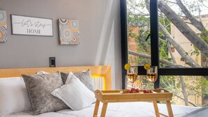 A bedroom with a bed, pillows, a bedside table with glasses and fruit, and a window with a view of trees and buildings. - Cozy Aparta-Suite Stadio| Rosa Negra (Medellín)