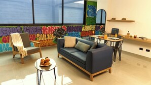 A modern living room with a sofa, armchair, and coffee table. There is a graffiti wall in the background and a window with blinds. - Cozy Aparta-Suite Stadio | Rosa Negra (Medellín)