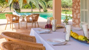 Un área de comedor al aire libre con sillas de mimbre, una mesa servida con platos, cubiertos y una bandeja de frutas.