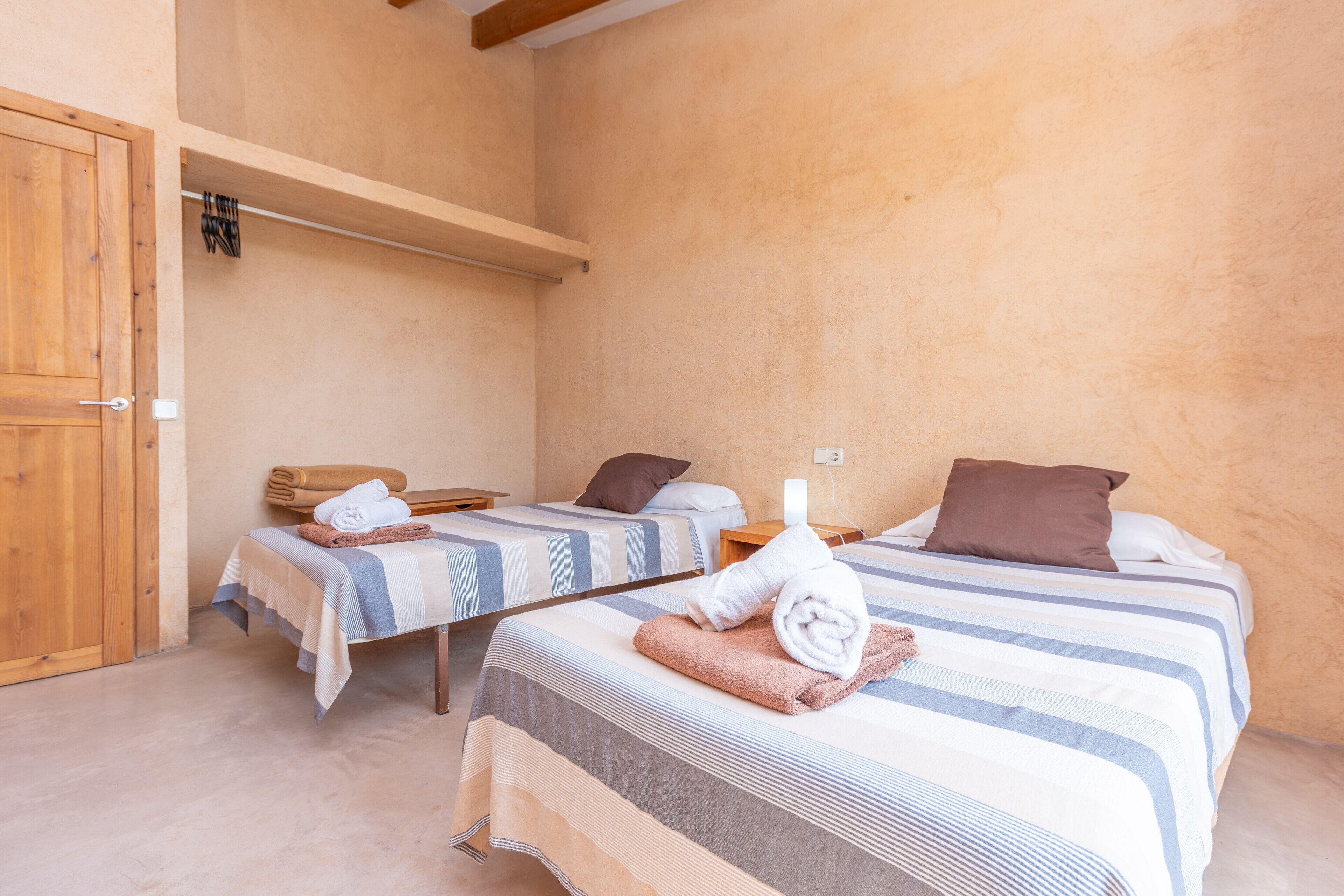 Habitación de hotel con dos camas, cada una con cubrecama y almohadas a rayas. Se aprecia una puerta de madera a la izquierda y un estante sobre las camas.