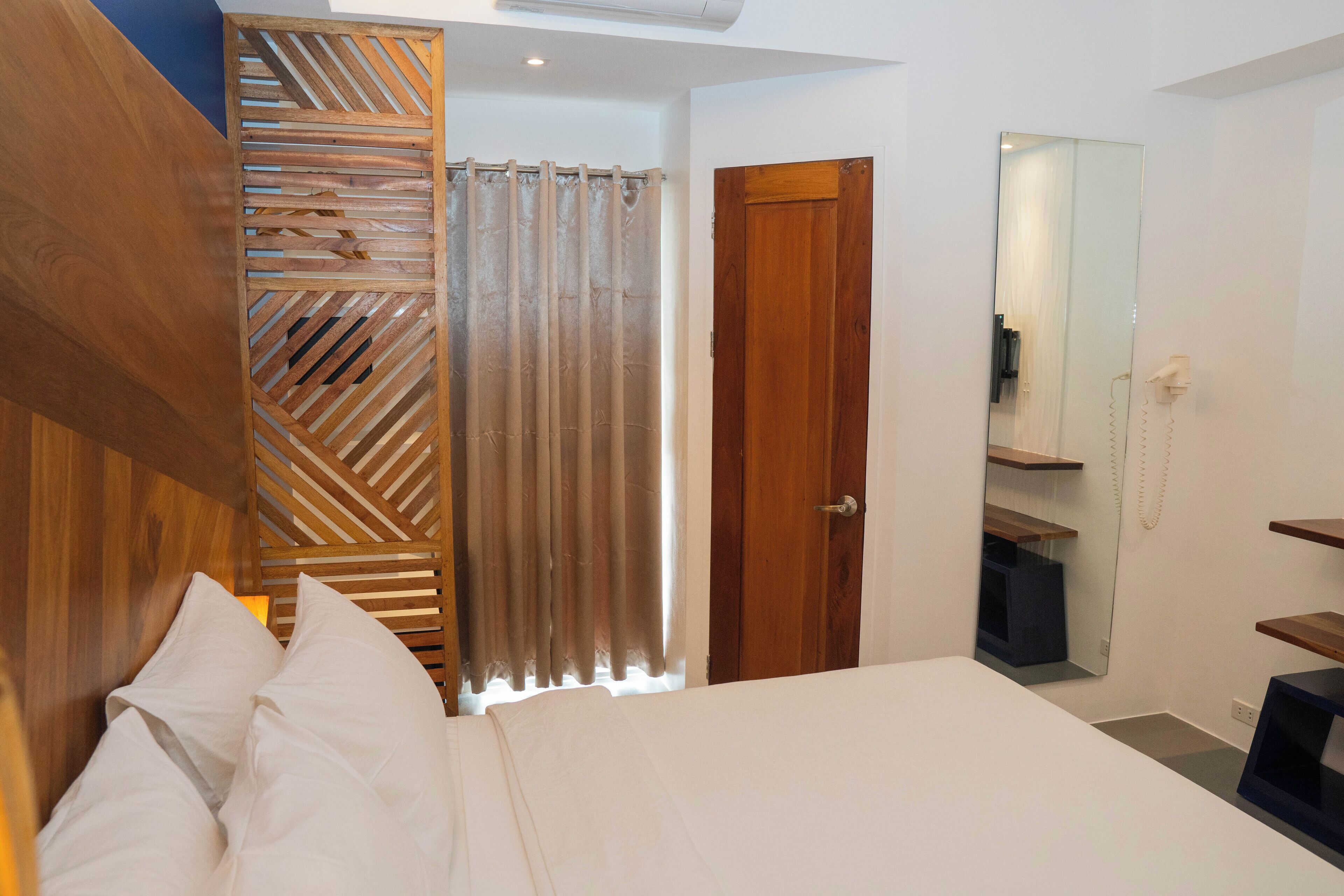 Quarto com cabeceira de madeira, cama com roupa de cama branca, porta de madeira, espelho e telefone.