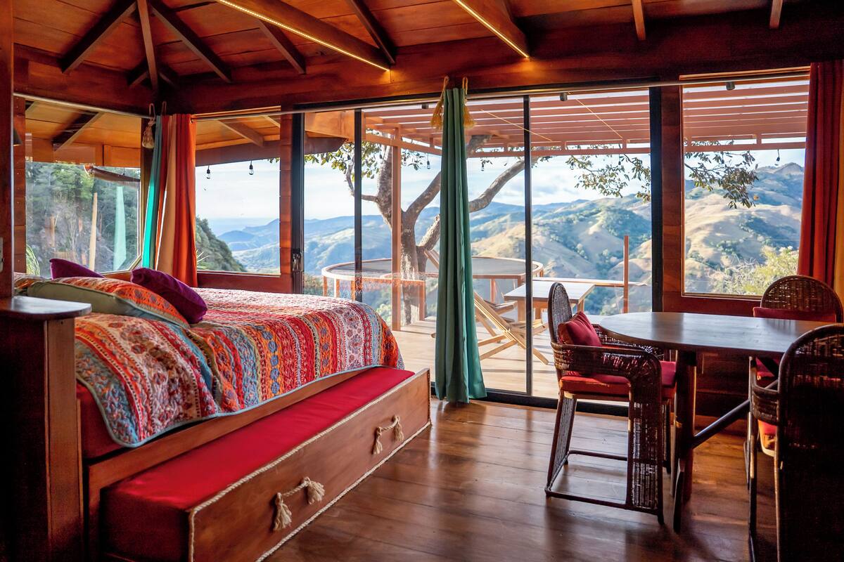 A wooden cabin with a bed, a dining table, and a view of the mountains.