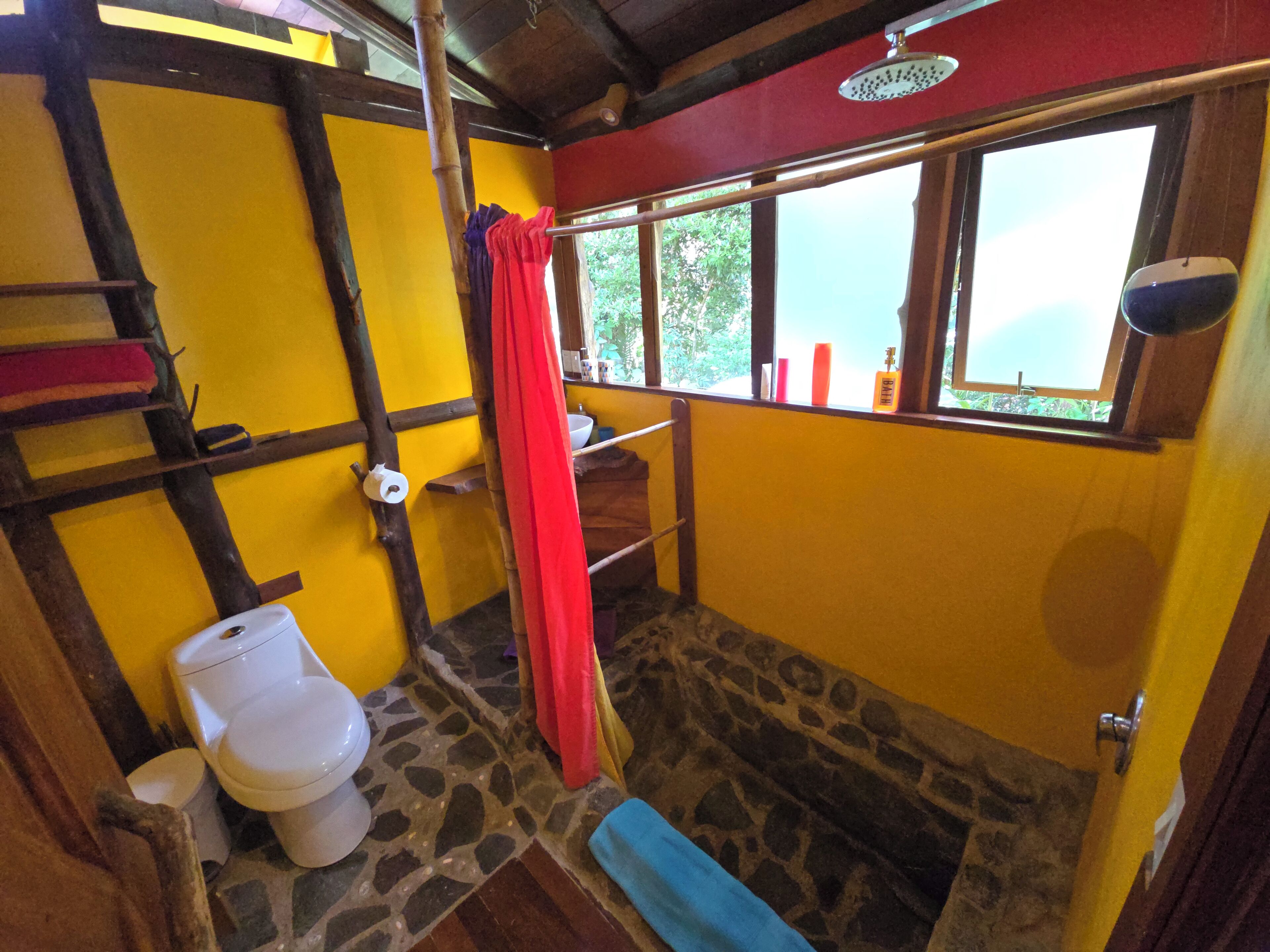 Un baño con una decoración en tonos amarillo y rojo, suelo de piedra, escalera de madera, inodoro, ducha y ventana con vistas a la vegetación.