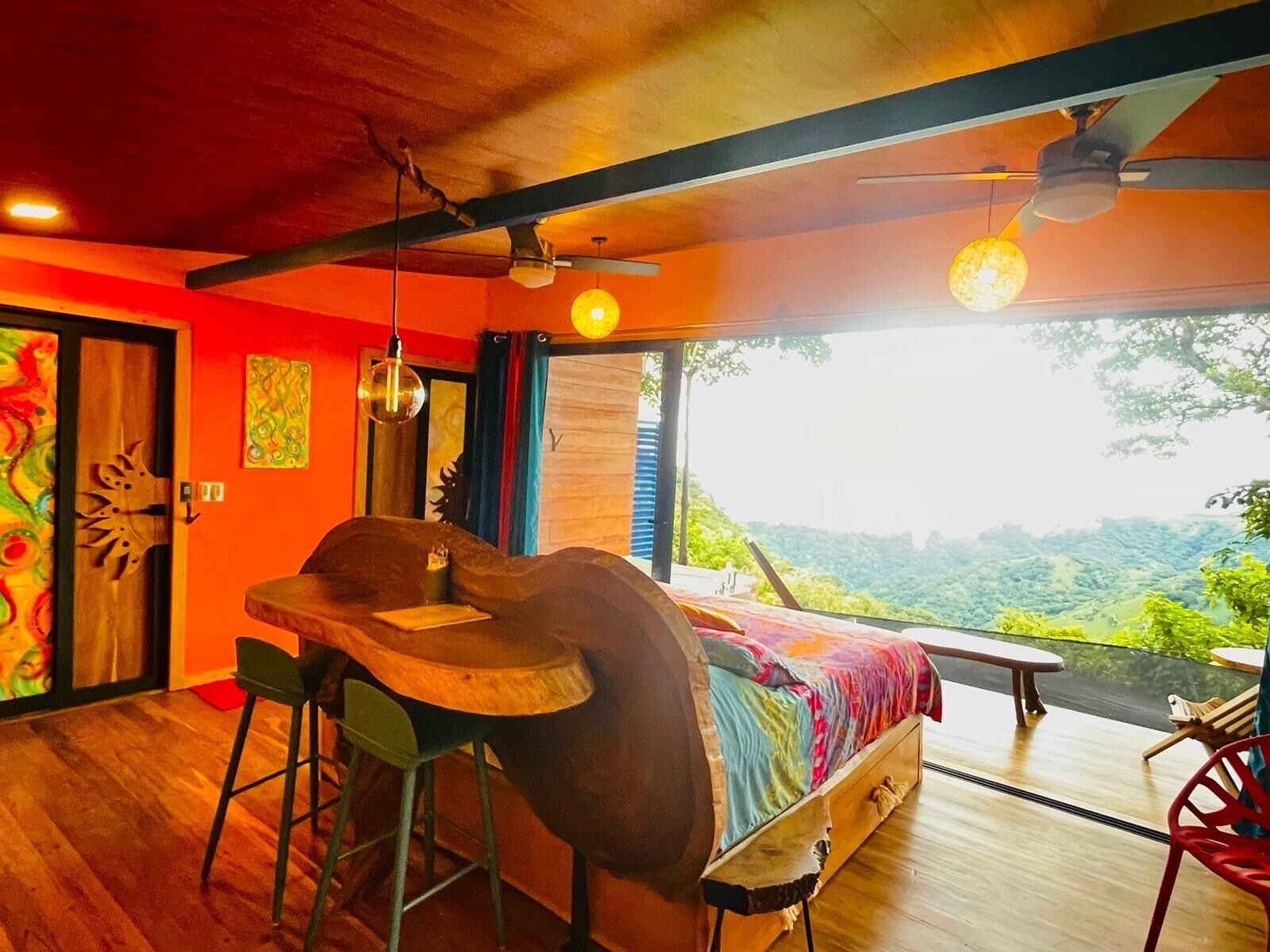 Una habitación con cama, una mesa de madera y una silla. Se ve un paisaje verde a través de la ventana.