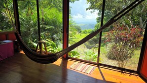 A hammock in a room with large windows overlooking a lush garden. - El Sol Monteverde (Monteverde)