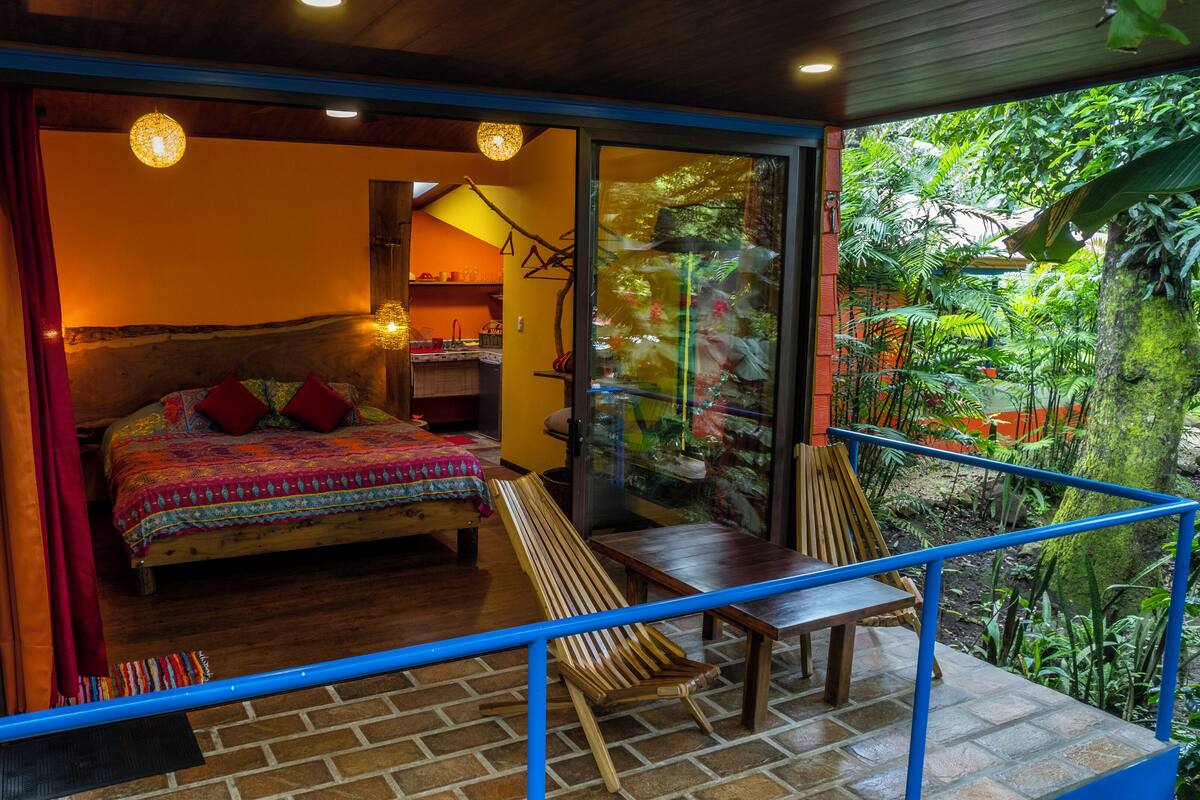 A bedroom with a bed, a wooden chair, a small table, and a glass door leading to a balcony with a view of greenery.