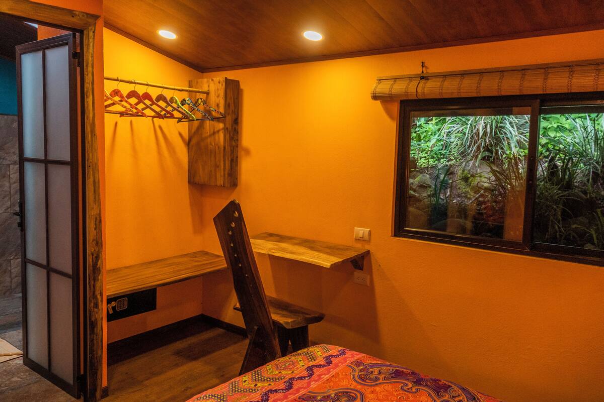 A room with a bed, a desk, a chair, a window with a view of greenery, and a wooden door.