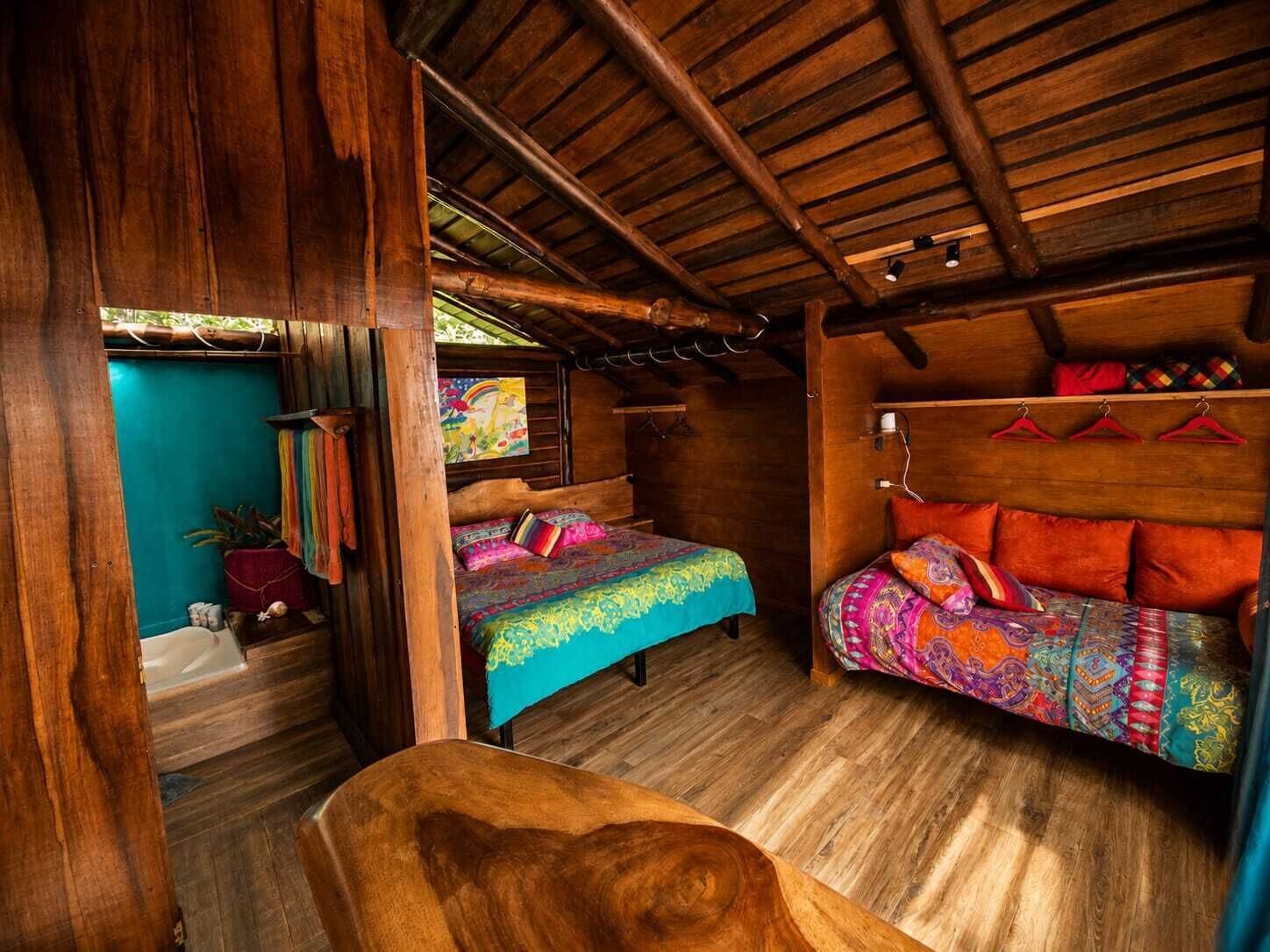 Interior de una cabaña de madera con una cama, una mesita y una hamaca.