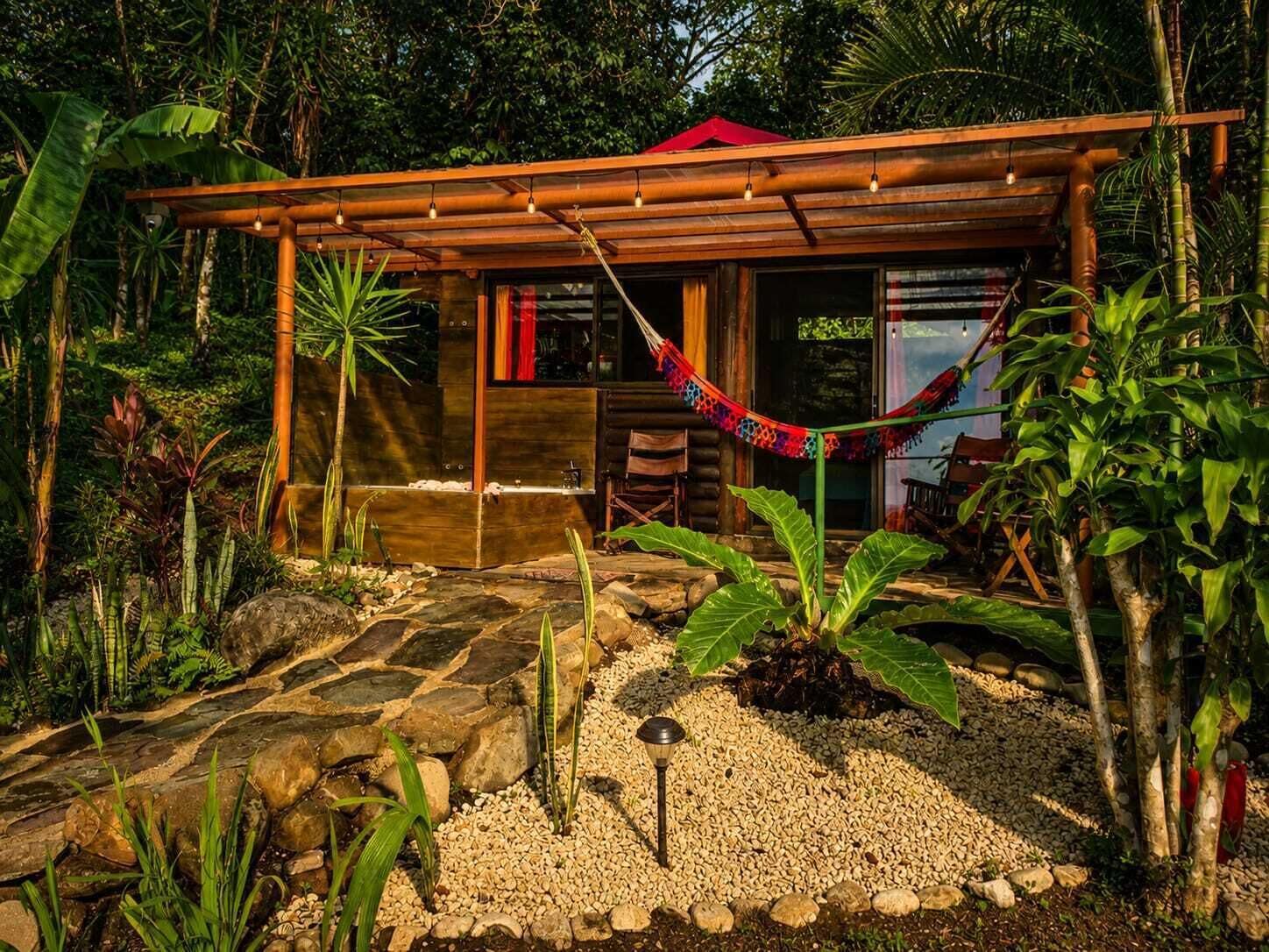 A wooden cabin with a hammock, surrounded by tropical vegetation and a stone pathway.
