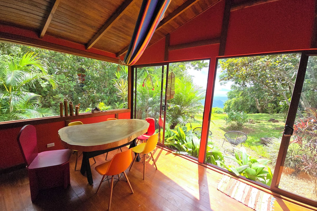 A room with a wooden table and chairs, large windows, and a view of greenery outside.