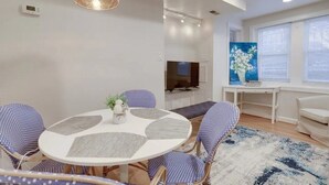 Una sala de estar moderna con una mesa de comedor redonda, sillas con estampado azul, un televisor de pantalla plana y un amplio ventanal con persianas.