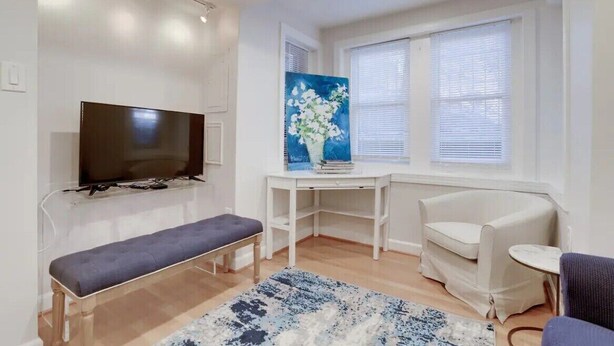 Una sala de estar moderna con un televisor de pantalla plana, un banco, una mesa blanca con un cuadro, un sillón blanco y una mesita redonda.