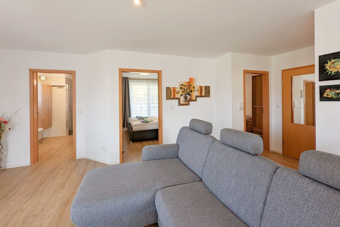 A modern living room with a grey sectional sofa, wooden flooring, and a hallway leading to a bedroom with a bed and a window.
