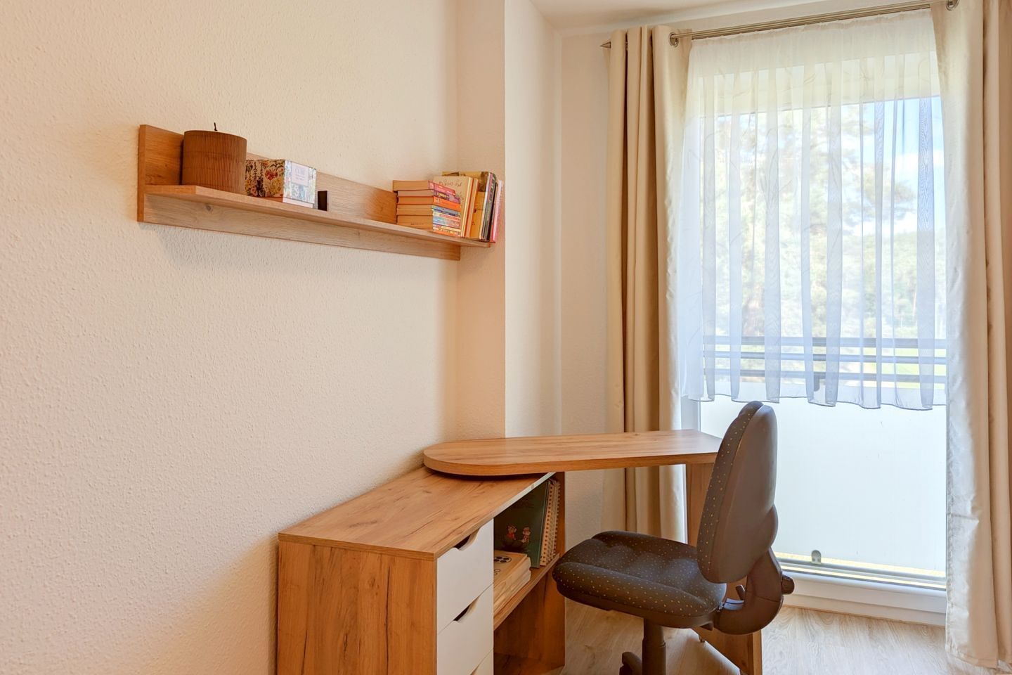 A small office space with a wooden desk, a chair, a shelf with books and a candle, and a window with curtains.