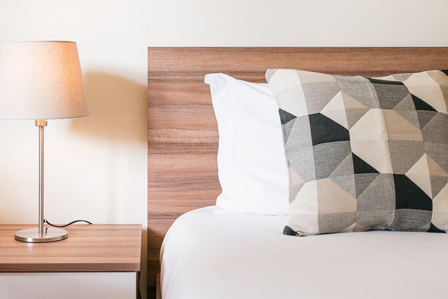 Una cama bien tendida con un cojín estampado con motivos geométricos y un cojín blanco liso, una lámpara de pie moderna y un cabecero de madera.
